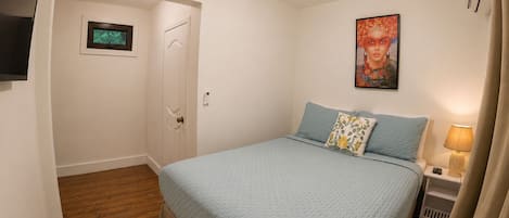 Ein Schlafzimmer mit einem Bett, einer Tür, einem an der Wand montierten Fernseher, einem Gemälde und einem Nachttisch mit einer Lampe.