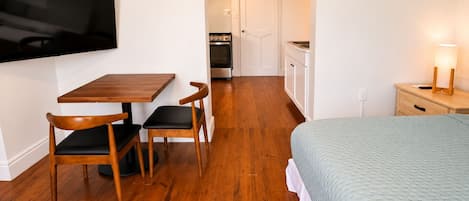 A room with a bed, a table with chairs, a TV mounted on the wall, a kitchen area with cabinets and a stove, and a bedside lamp.