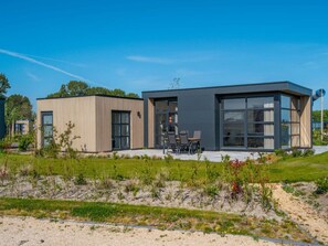 Een modern gebouw met twee verdiepingen, grote ramen, een terras met buitenmeubilair en een verzorgde tuin.