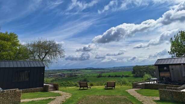 Ländliche Landschaft mit blauem Himmel, zwei Holzhütten und einer Bank im Vordergrund.