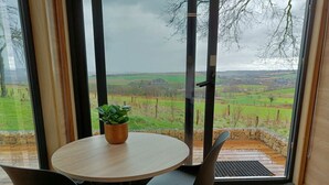 Ein Zimmer mit einem runden Tisch, zwei Stühlen, einer Topfpflanze und Blick auf eine grüne Landschaft durch große Fenster.