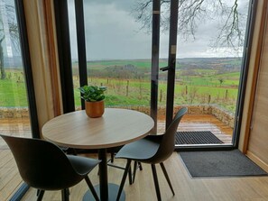 Ein Zimmer mit einem runden Tisch, zwei Stühlen, einer Topfpflanze und Blick auf eine grüne Landschaft durch große Fenster.