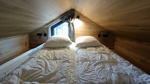 Ein kleiner, gemütlicher Raum mit schrägem Dach, einem Einzelbett mit zwei Kissen, einem Fenster mit Vorhängen und einer Wandleuchte.