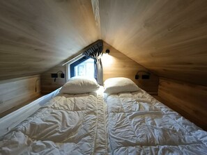 Ein kleiner, gemütlicher Raum mit schrägem Dach, einem Einzelbett mit zwei Kissen, einem Fenster mit Vorhängen und einer Wandleuchte.