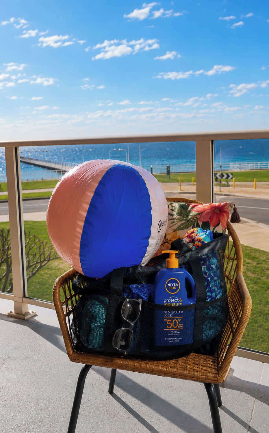 A wicker chair with a beach ball, a blue sunscreen bottle, and a black bag on a balcony with a view of the sea and a bridge.