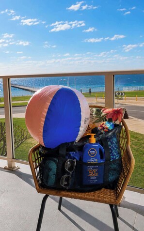 A wicker chair with a beach ball, a blue sunscreen bottle, and a black bag on a balcony with a view of the sea and a bridge. - Rockingham Retreat | Right By The Beach (Rockingham)
