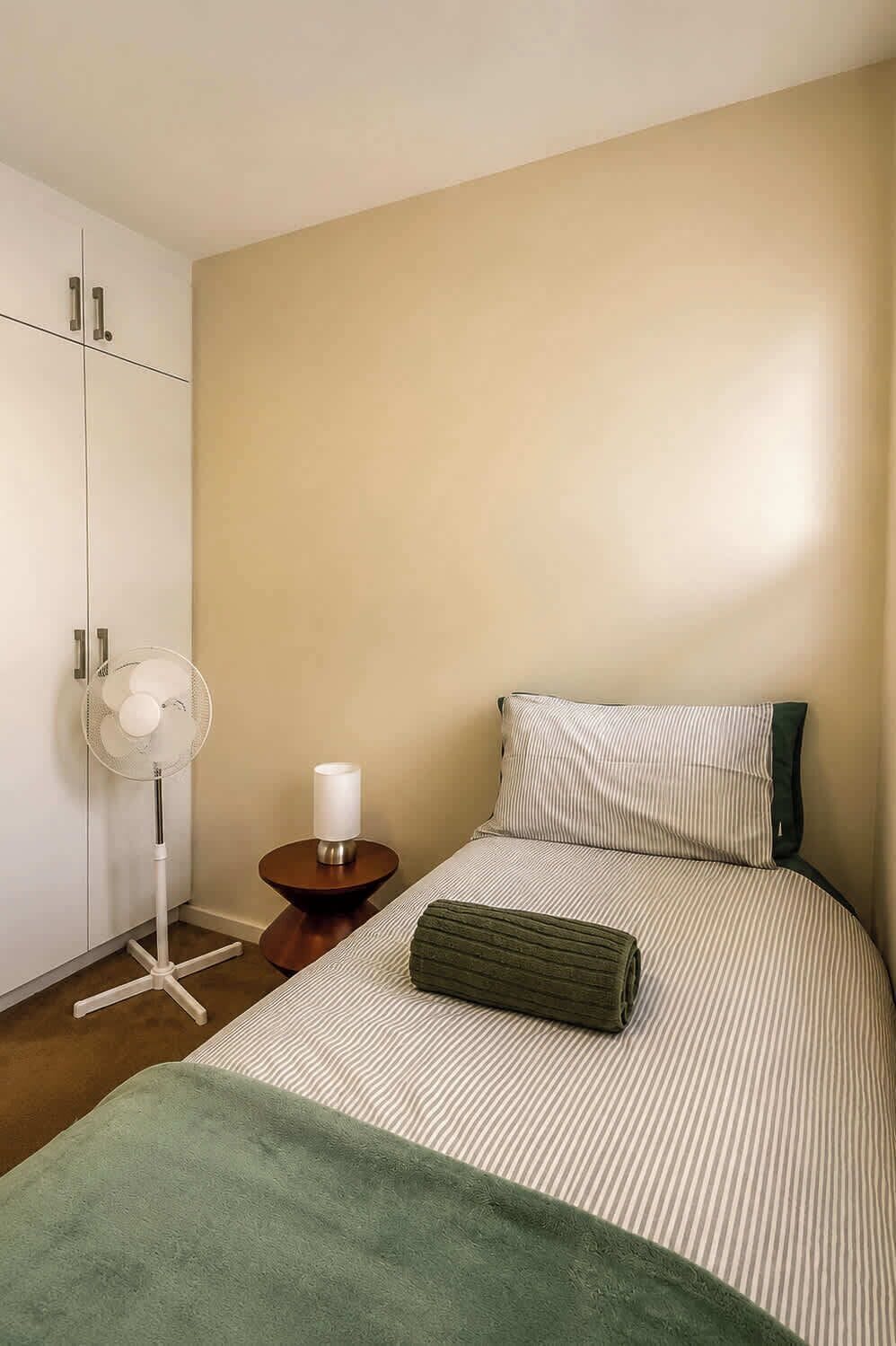 A bedroom with a bed, a bedside table with a lamp, a standing fan, and a wardrobe.
