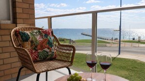 A wicker chair with a floral cushion, a round wooden table with a cheese board, grapes, and wine glasses, overlooking a body of water. - Rockingham Retreat | Right By The Beach (Rockingham)