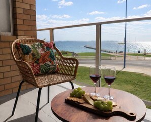 A wicker chair with a floral cushion, a round wooden table with a cheese board, grapes, and wine glasses, overlooking a body of water. - Rockingham Retreat | Right By The Beach (Rockingham)