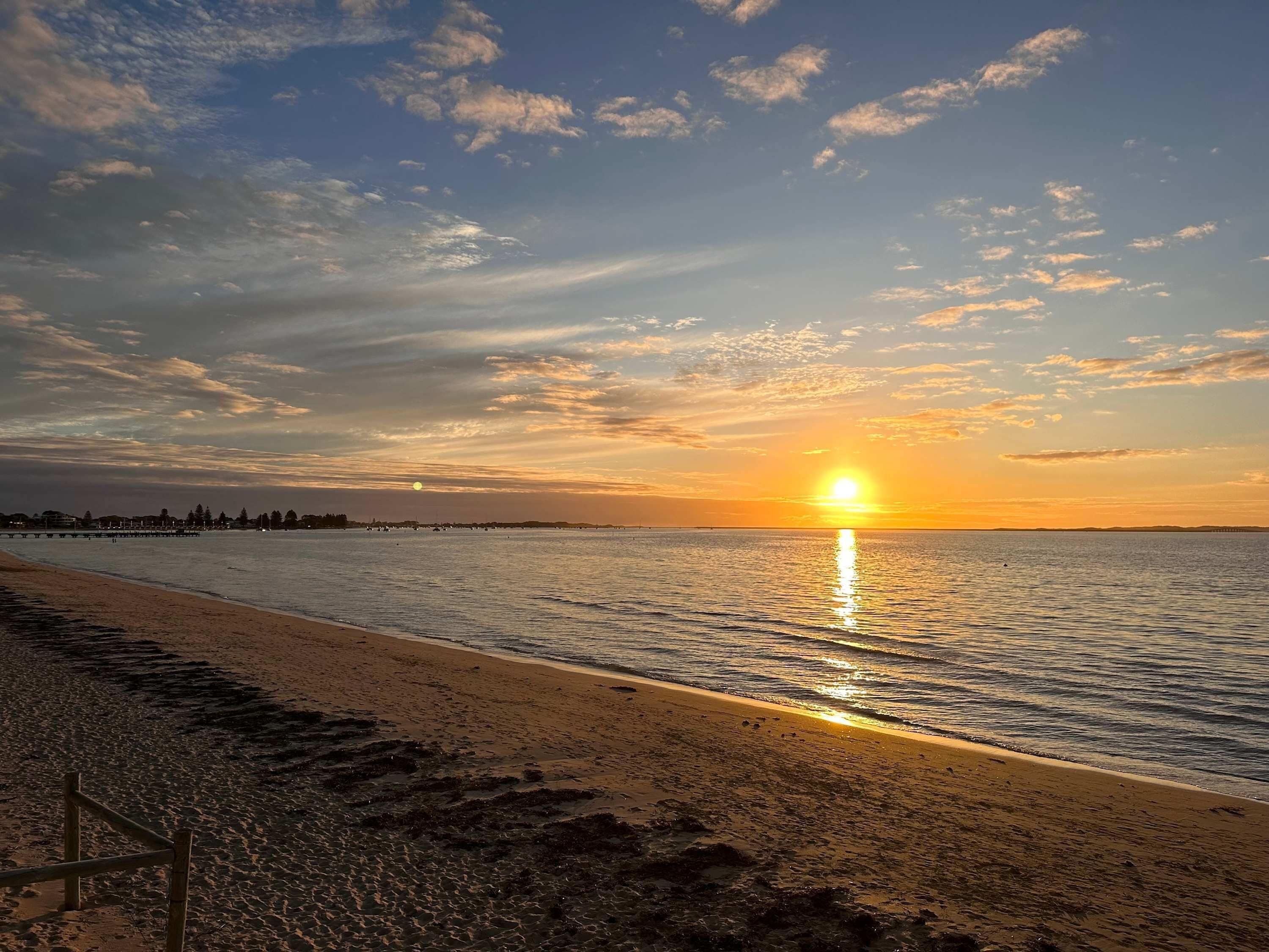A beach with a sunset, calm water, and a pier in the distance.
