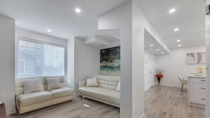 A modern living room with a beige sofa, a dining area with a white table and chairs, and a painting on the wall.