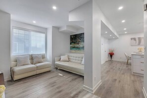 A modern living room with a beige sofa, a dining area with a white table and chairs, and a painting on the wall.