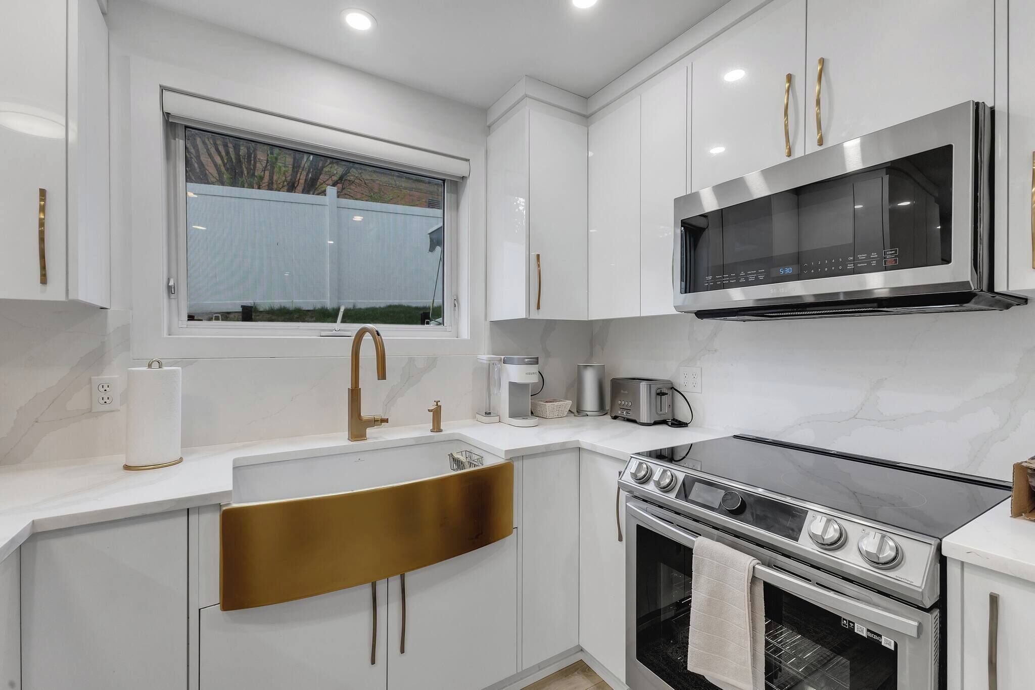 Une cuisine moderne avec des meubles blancs, un four à micro-ondes en acier inoxydable, une cuisinière à gaz et une fenêtre avec des stores.