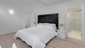 Un dormitorio moderno con una cama grande, lámparas de noche, una mesita de noche y un vestidor con puerta de cristal.