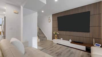 A modern living room with a large flat-screen TV mounted on a wooden panel wall, a sofa, a coffee table, and a staircase in the background.