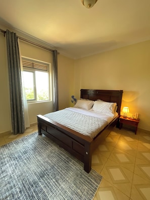 Una camera da letto con un letto, un comodino, una lampada, una finestra con tende e un tappeto decorato.
