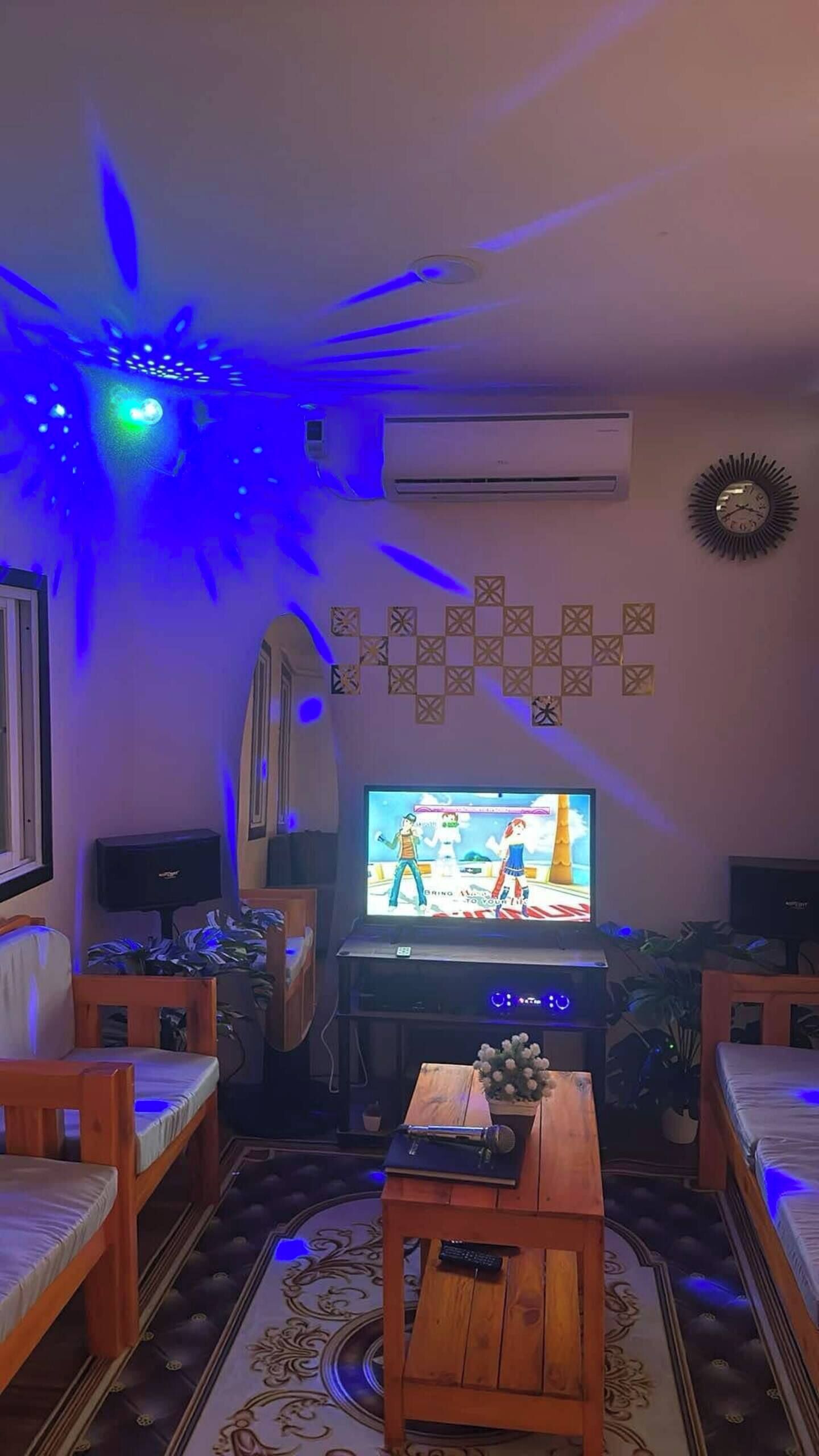 A living room with a television, wooden furniture, decorative wall tiles, and a ceiling light fixture.