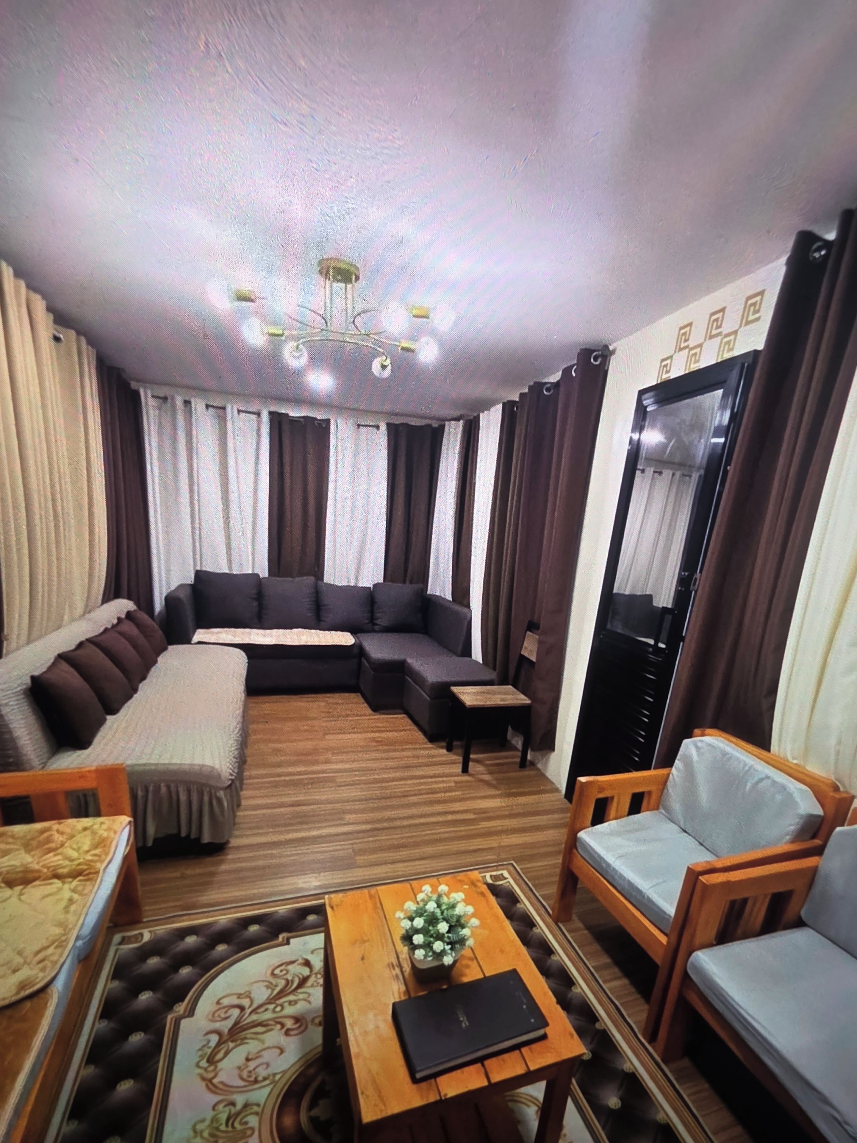 Un salon comprenant un canapé d’angle, une table basse en bois avec un livre et des fleurs, une petite chaise en bois et un grand miroir accroché au mur.