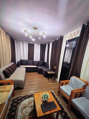 A living room with a sectional sofa, a wooden coffee table with a book and flowers, a small wooden chair, and a large mirror on the wall.