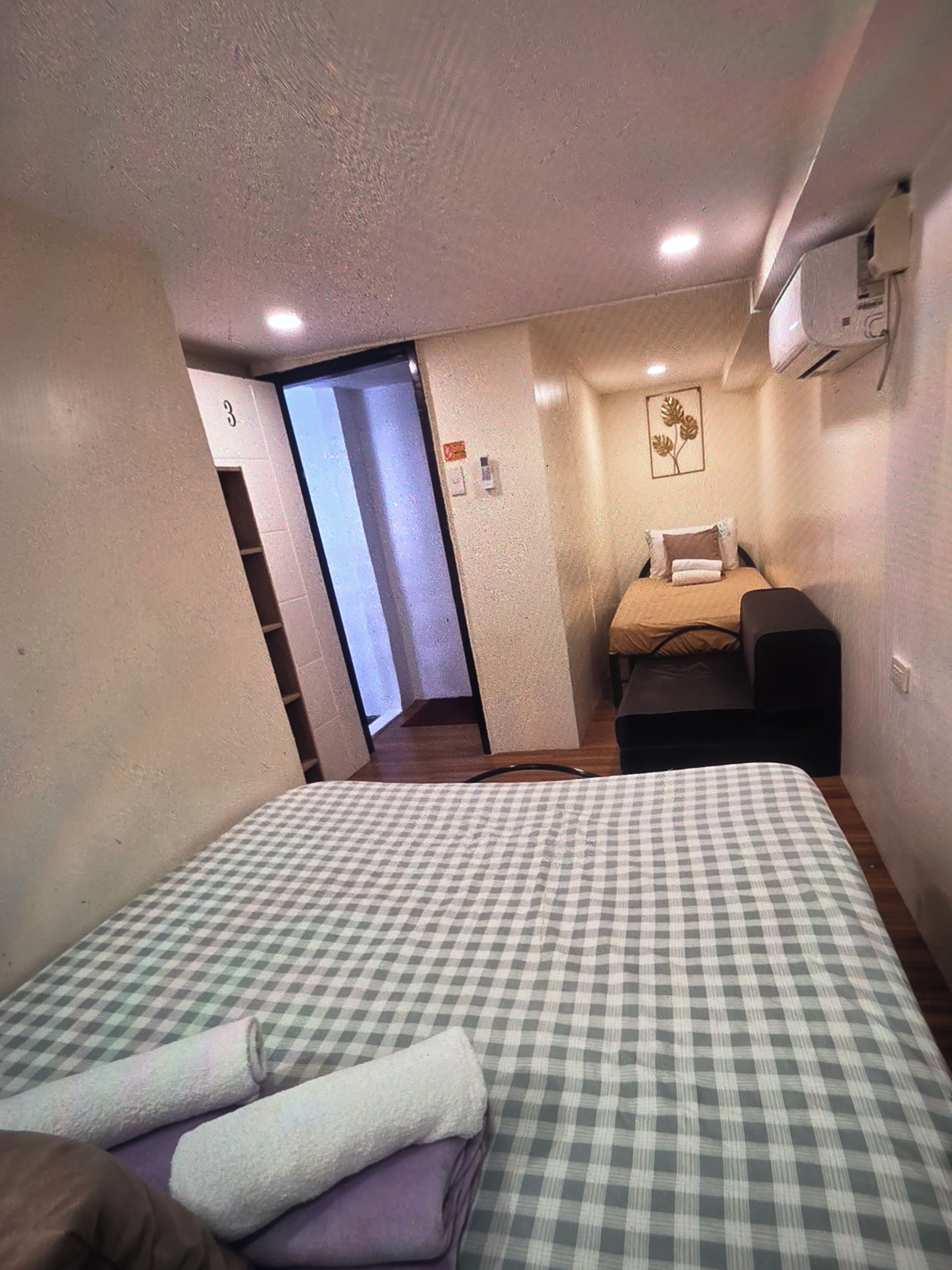 A hotel room with a checkered bedspread, a bedside table, a chair, and a small desk. There is a door leading to another room and an air conditioning unit mounted on the wall.