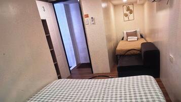 A hotel room with a checkered bedspread, a bedside table, a chair, and a small desk. There is a door leading to another room and an air conditioning unit mounted on the wall.