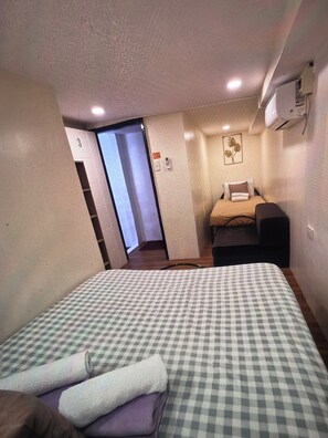 A hotel room with a checkered bedspread, a bedside table, a chair, and a small desk. There is a door leading to another room and an air conditioning unit mounted on the wall.