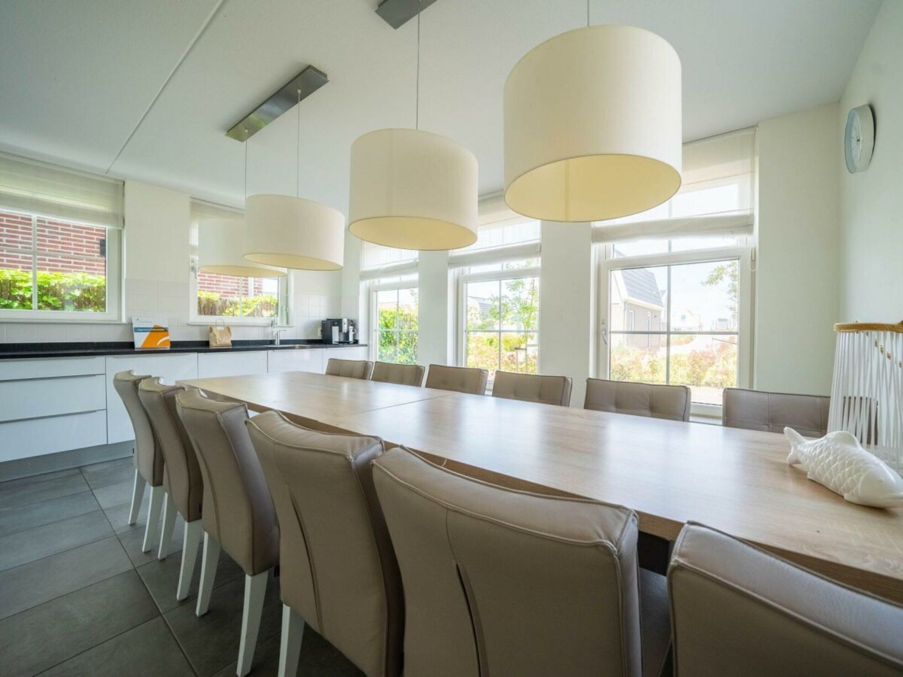 A modern kitchen with a long dining table, beige chairs, pendant lights, and large windows.