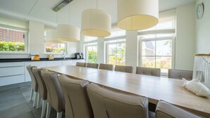 A modern kitchen with a long dining table, beige chairs, pendant lights, and large windows. - Holiday Home in Uitdam With Terrace (Uitdam)