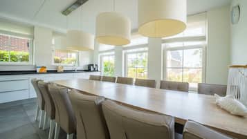 A modern kitchen with a long dining table, beige chairs, pendant lights, and large windows.