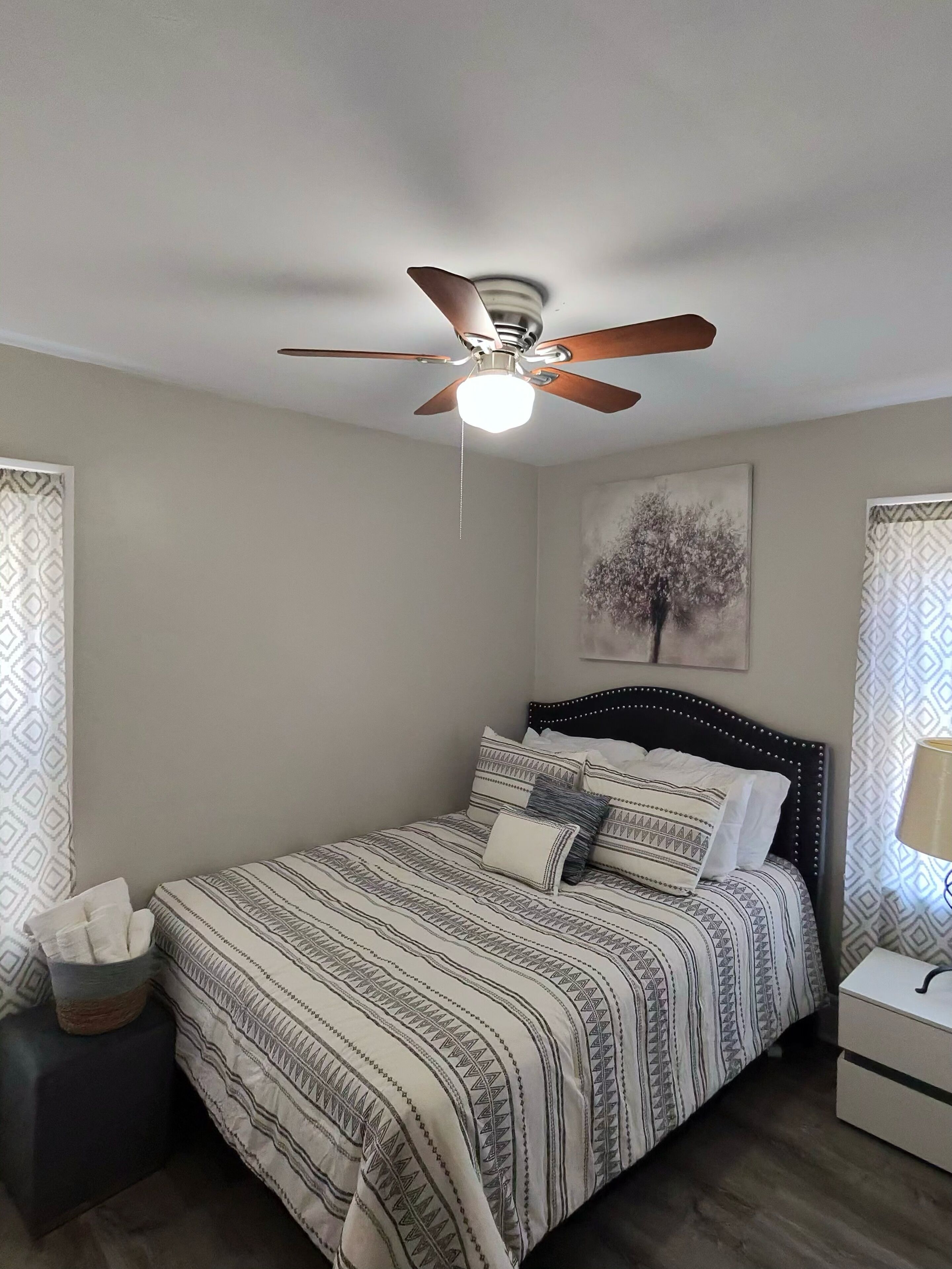 Une chambre à coucher comprenant un lit, un ventilateur de plafond, une table de chevet, une lampe et un mur orné d’un tableau encadré.