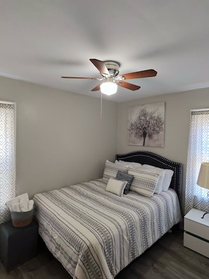 A bedroom with a bed, a ceiling fan, a nightstand, a lamp, and a wall with a framed picture.