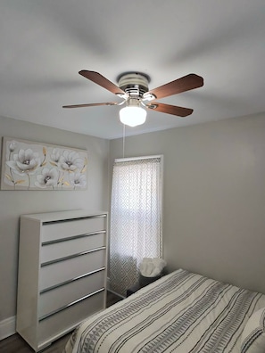 A bedroom with a ceiling fan, a bed with a patterned bedspread, a dresser with drawers, and a wall with a floral painting.