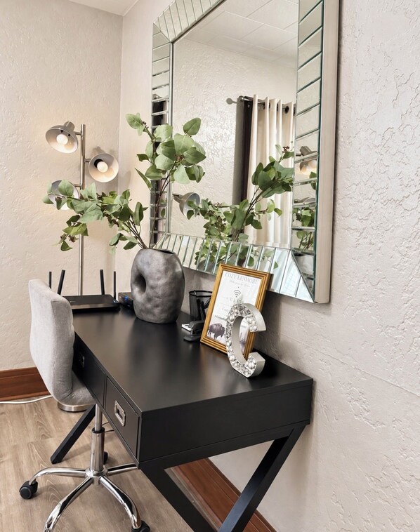 Een zwart bureau met een stoel, een vaas met groene planten, een ingelijste foto en een spiegel aan de muur.
