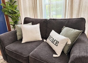 A dark grey sofa with three cushions, one with 'HOME' text, in a room with a plant and a window with curtains.