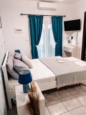 Ein Schlafzimmer mit einem Bett, einem Nachttisch mit Lampe, einem an der Wand montierten Fernseher und einem Fenster mit Vorhängen.