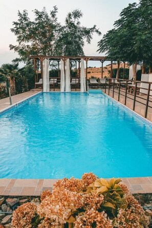 Una piscina rectangular con agua azul cristalina, rodeada por un solárium con baldosas y una estructura de pérgola.