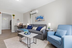 Ein Wohnzimmer mit einem grauen Sofa, einem blauen Sessel, einem weißen Couchtisch und einem Baumgemälde an der Wand.