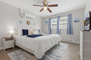 Ein Schlafzimmer mit Bett, Nachttisch, Deckenventilator, Klimaanlage, Fenster mit Vorhängen und einem Kunstwerk an der Wand.