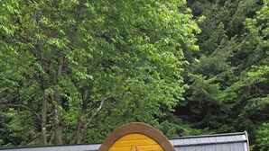 A small, yellow wooden building with a curved roof, a glass door, and a sign reading 'FLOKE'. There are two metal chairs and a small table in front.