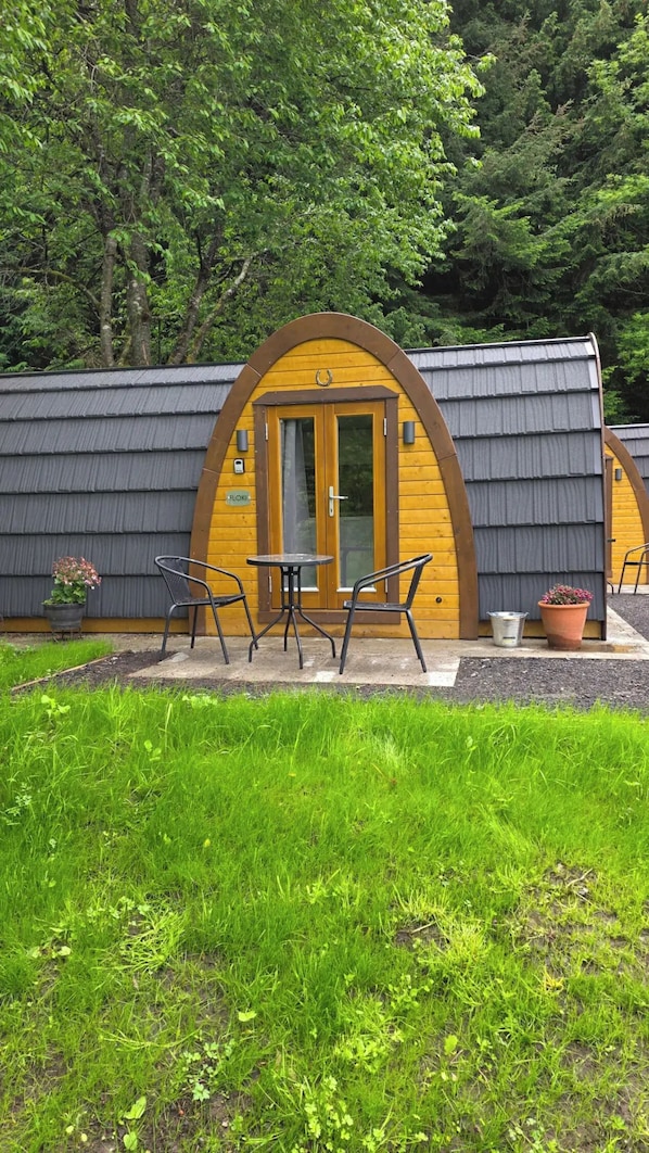 A small, yellow wooden building with a curved roof, a glass door, and a sign reading 'FLOKE'. There are two metal chairs and a small table in front.
