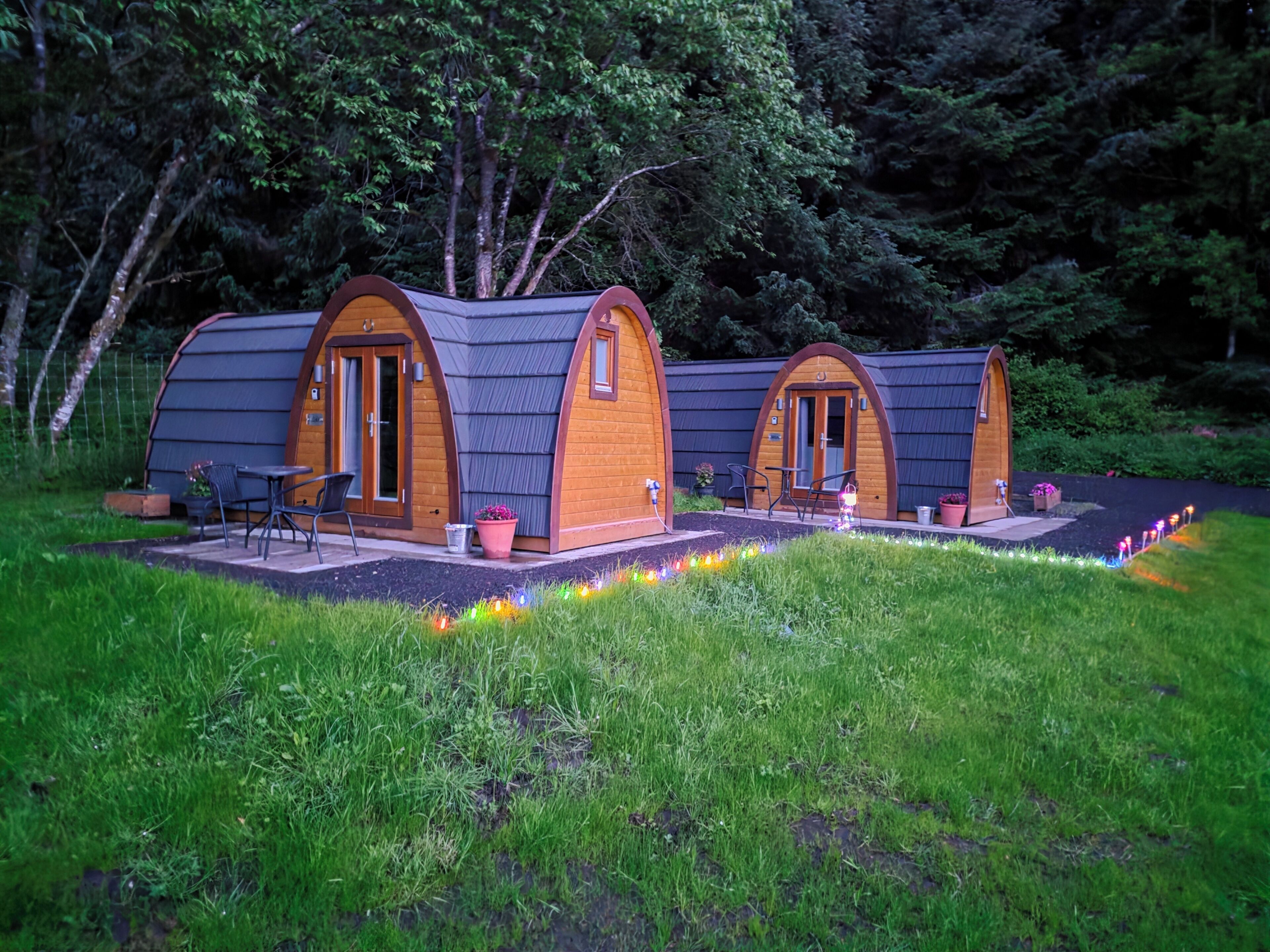 Two wooden A-frame cabins with doors and windows, surrounded by a grassy area and trees.