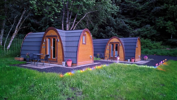 Two wooden A-frame cabins with doors and windows, surrounded by a grassy area and trees.