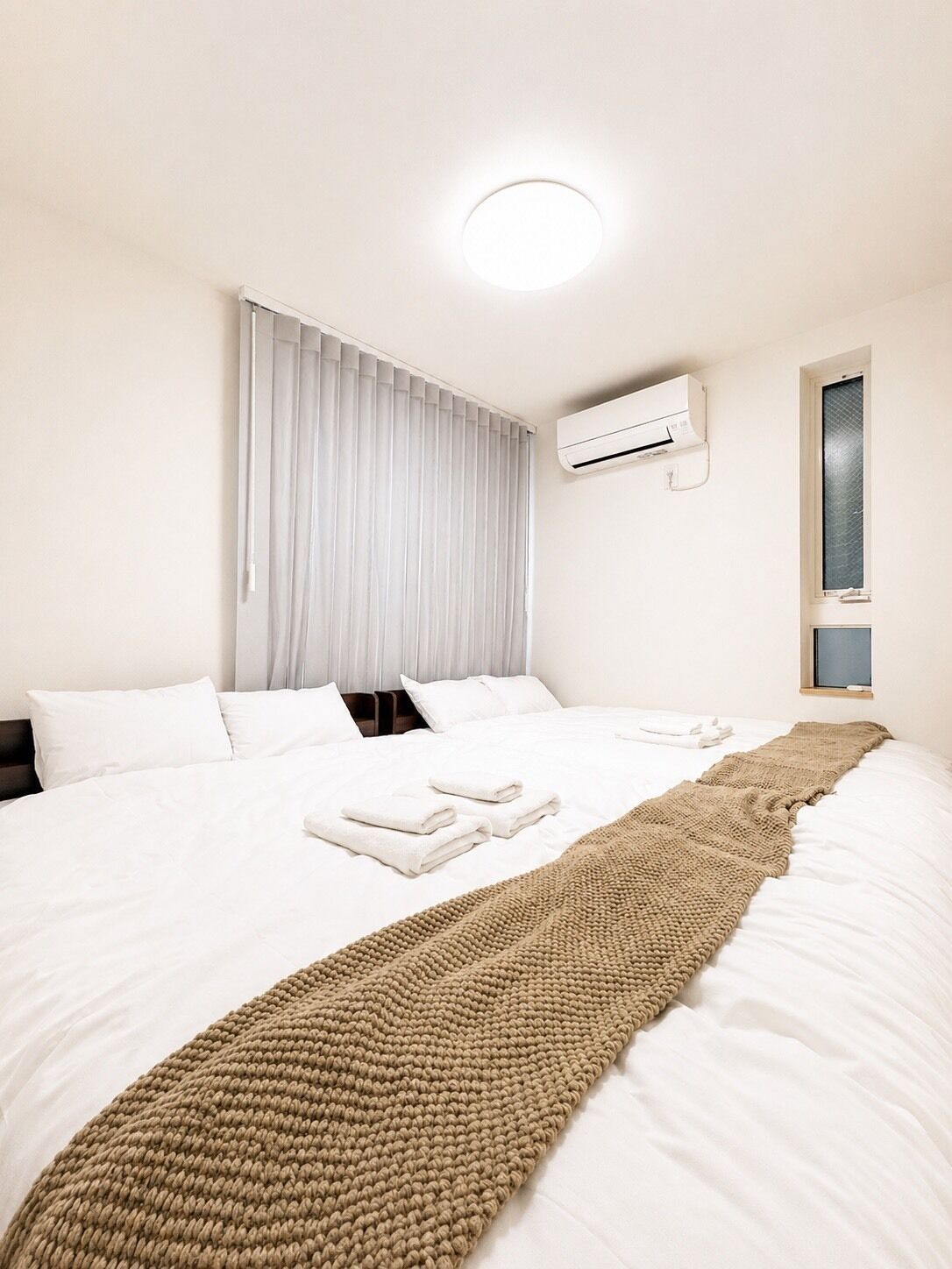 A hotel room with a large bed, white bedding, a beige throw blanket, an air conditioning unit, and a window with blinds.