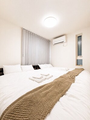 A hotel room with a large bed, white bedding, a beige throw blanket, an air conditioning unit, and a window with blinds. - 7-minute walk from Iriya Station | Accommodates up to 12 guests | Private rental: 111 ㎡ | 2-minute walk to a 24-hour supermarket (台東区)