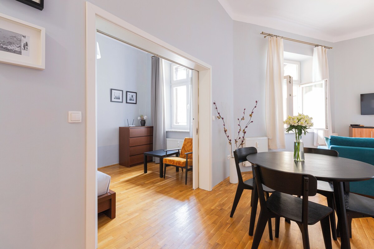A modern living room with a dining area, wooden flooring, and a large window with curtains.