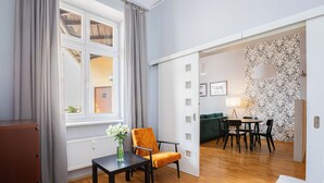 Et værelse med trægulv, et bord med stol, en radiator, et vindue med gardiner og en dør til et andet værelse.
