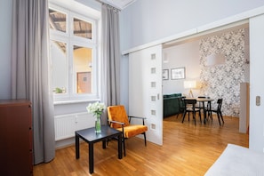 A room with a wooden floor, a table with a chair, a radiator, a window with curtains, and a door leading to another room. - Native Apartments Łazarza 3 (Kraków)