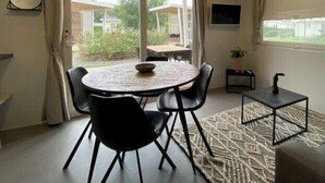 Een kamer met een ronde tafel, vier stoelen, een klein bijzettafeltje, een televisie en een raam met gordijnen.