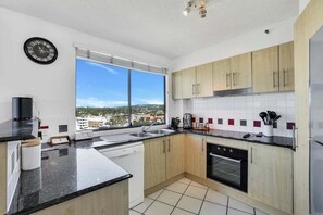 A modern kitchen with a dishwasher, oven, and a large window offering a city view. - Amazing Ocean Views from Unit 87-Catalina-II (Maroochydore)