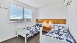 Une chambre avec deux lits simples, une fenêtre avec des stores, une table de chevet avec des lampes et un appareil de climatisation.